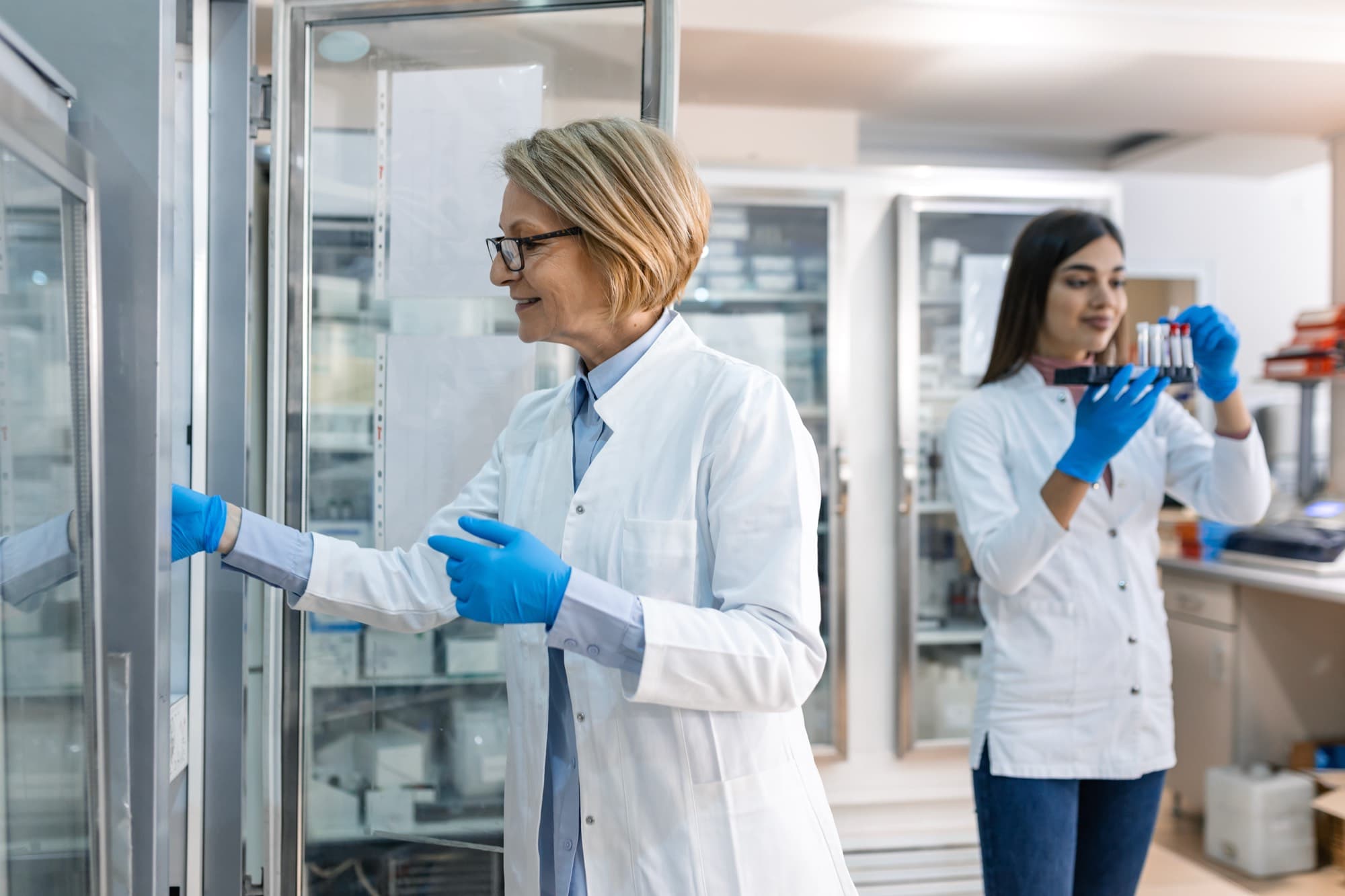 Two scientists in white coats and blue gloves at a laboratory's glass-door cold storage cabinets, one opening a cabinet and one holding a rack of sample tubes